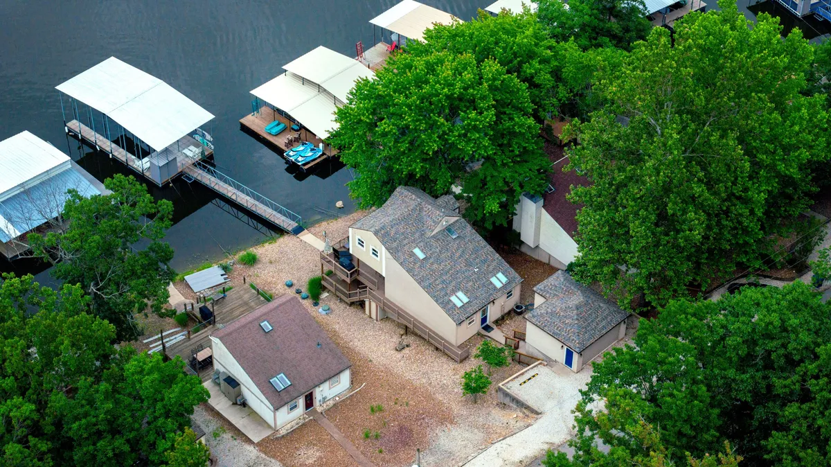 A wide exterior view of the main house, its boathouse, multiple docks, and lush surrounding trees. The scene shows the property’s direct proximity to the water and the ample dock space for boats and watercraft.