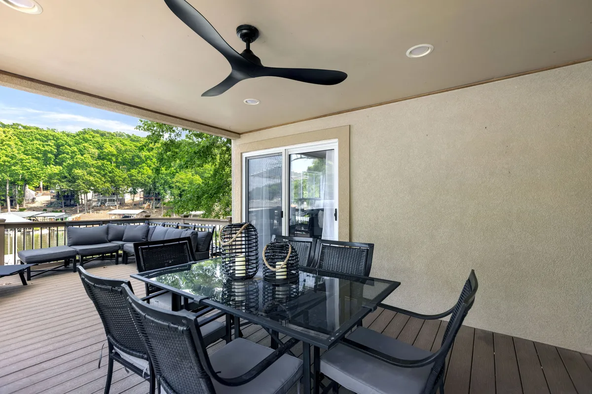 A covered outdoor dining area featuring a glass dining table surrounded by black wicker chairs, a ceiling fan for comfort, and an outdoor sofa. The space is styled for al fresco dining and relaxation, with views extending beyond the patio.