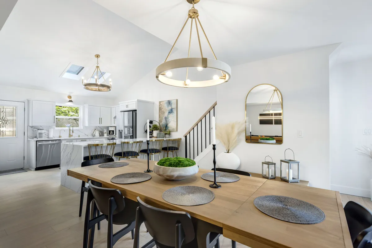 This image showcases a large dining table beneath a glowing chandelier, with a modern kitchen counter visible in the background and tasteful artwork on the wall. The space is set up for family dinners, entertaining guests, and culinary activity.