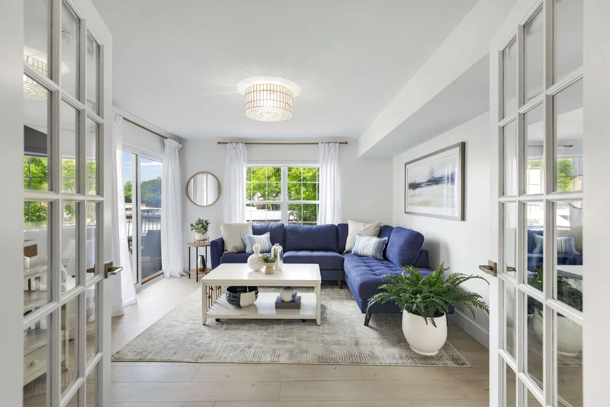 A serene living space with a large blue sectional sofa, coffee table, and a picture window allowing in an abundance of natural light. The view through the window hints at the lake, making the water a subtle but constant presence within the home.