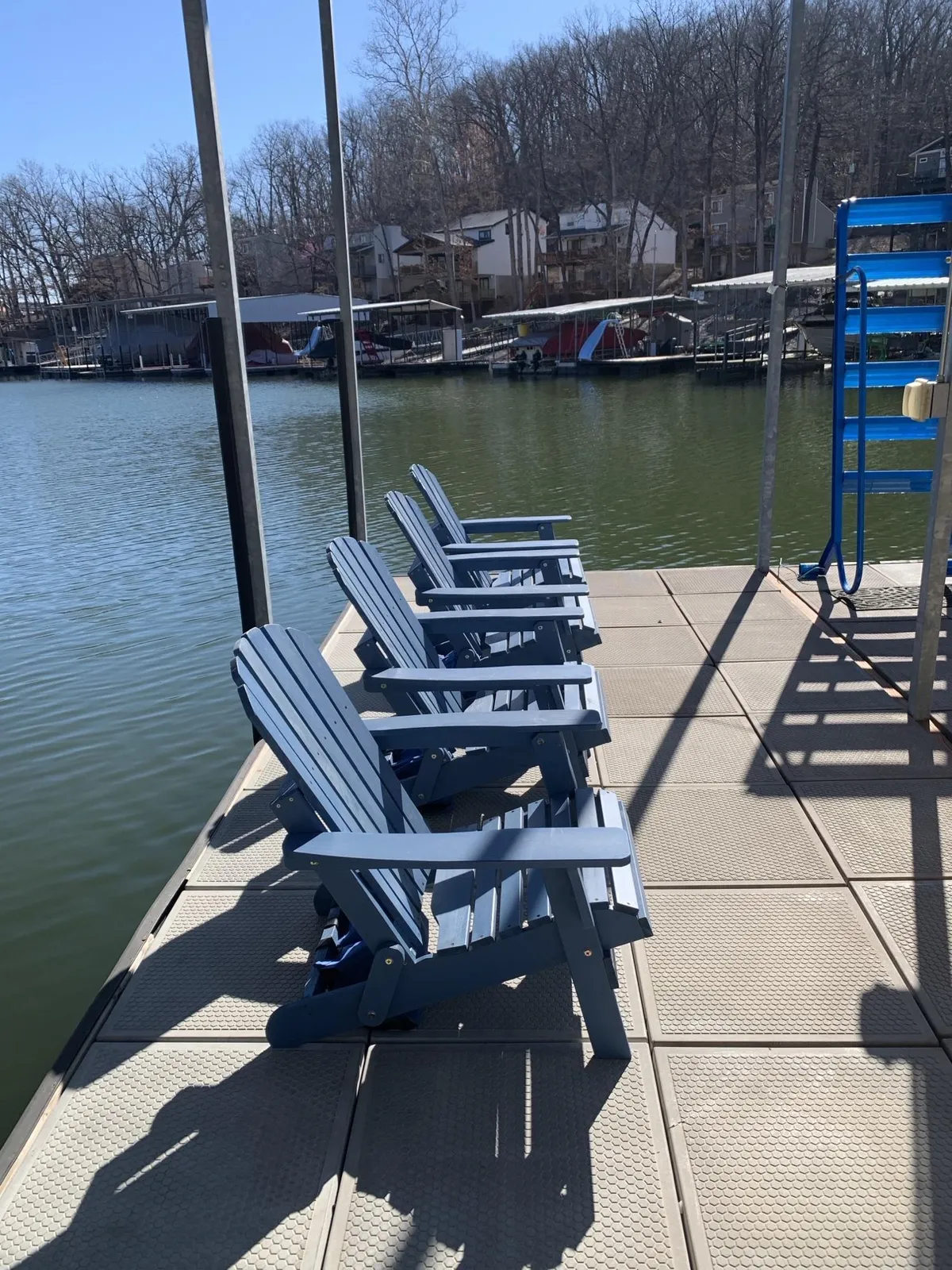 The image features blue Adirondack chairs positioned right by the calm water, with docks and boats visible in the background, suggesting community and lake activity. The tranquil setting suggests both solitude and the potential for social gatherings by the lake.