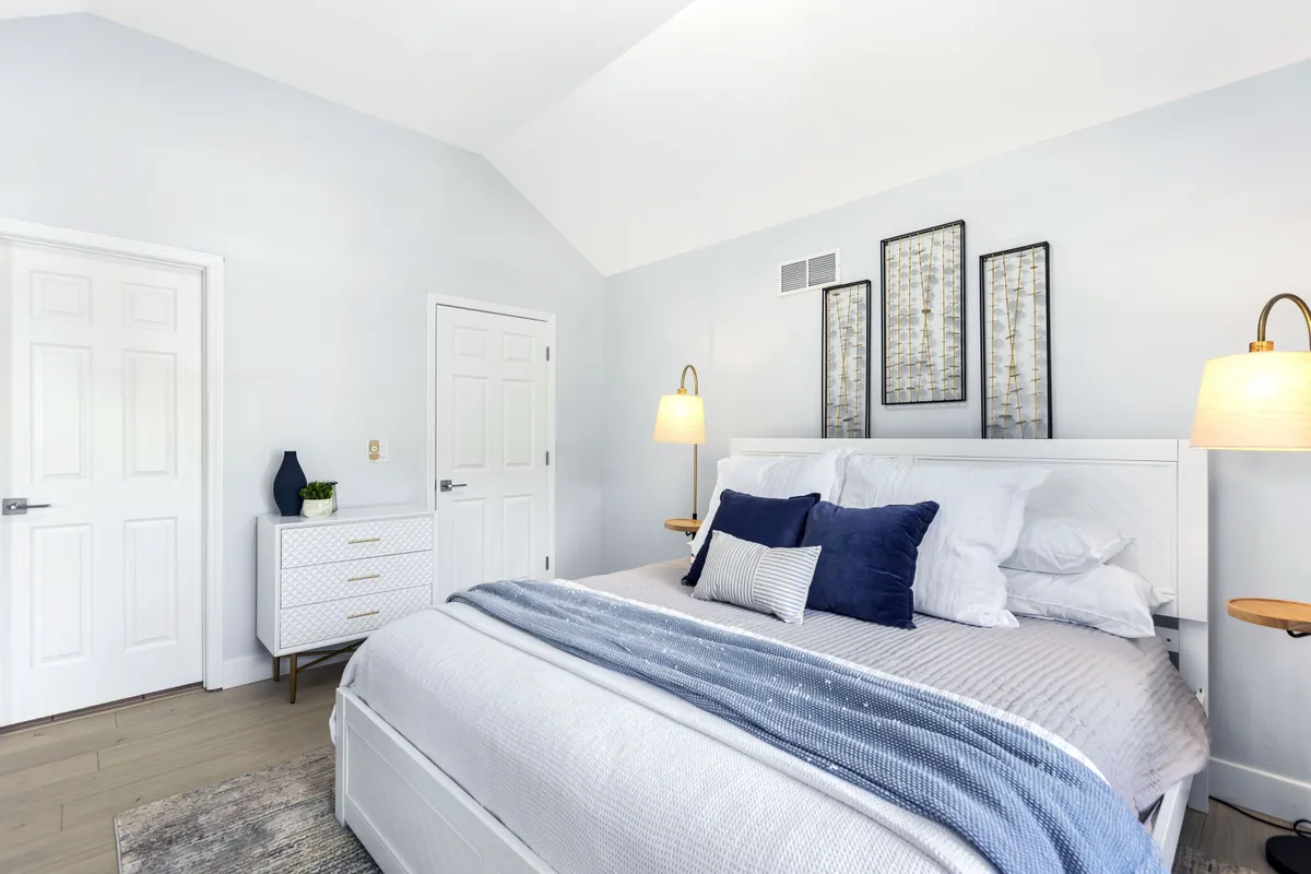 A stylish bedroom anchored by a large bed with plush, decorative pillows, complemented by serene wall art and functional nightstands with soft-glow lamps. A dresser stands nearby, and natural light floods the room, creating a calm, restful environment ideal for relaxation or reading.