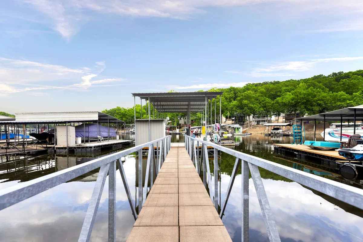 A scenic walkway leads to the dock, where boats are moored against a backdrop of trees and calm water. The space is inviting for both adults and kids to start their lakeside explorations, whether it's for launching paddle boards or fishing at sunrise.