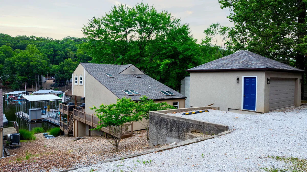 An exterior shot of the house, garage, dock area, and surrounding trees. The image presents the tranquil lakeside environment, with easy access to the water and ample space for relaxation or social gatherings outdoors.