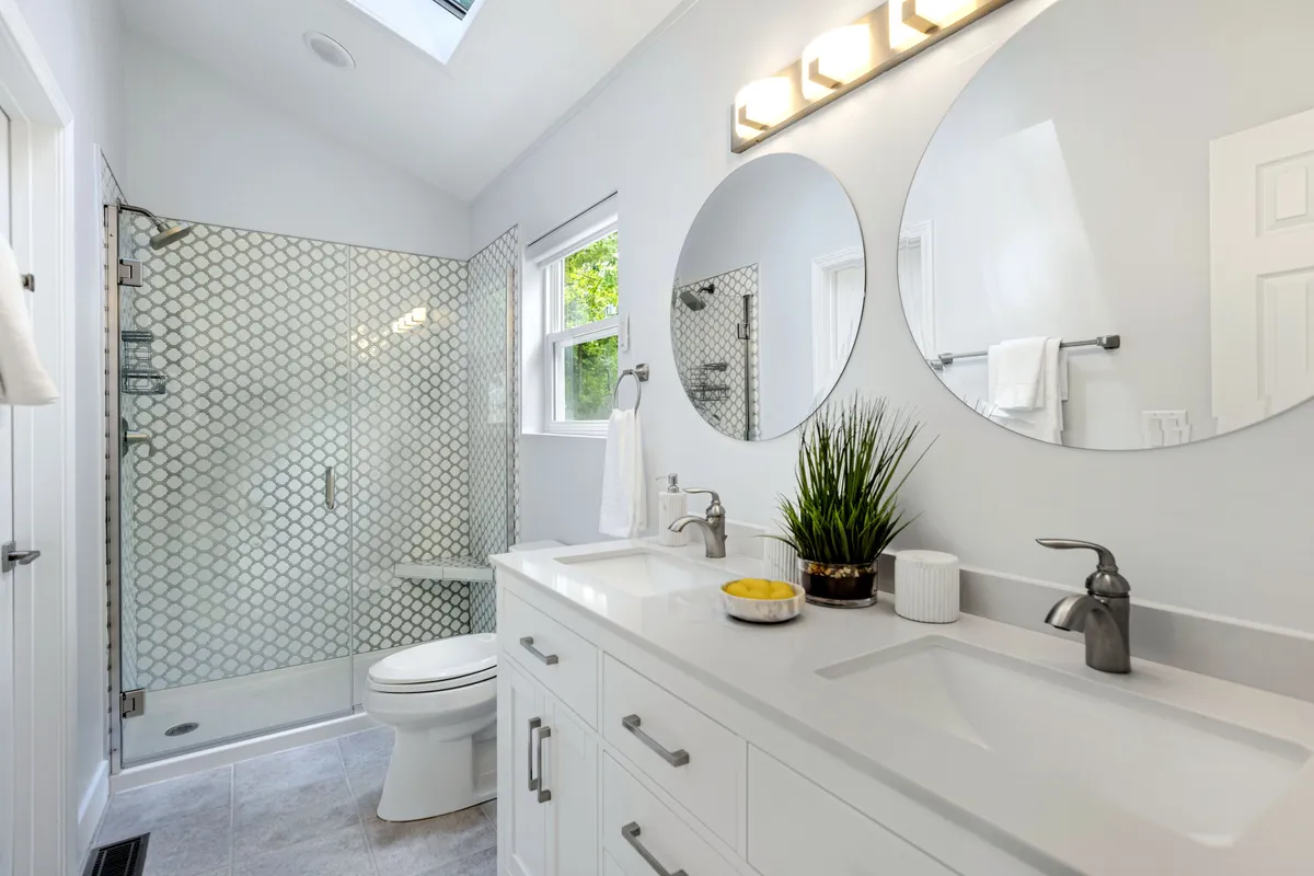 A modern and elegant bathroom with a double sink vanity, expansive mirrors, a glass shower enclosure, and decorative plants. The space is flooded with soft light, evoking a tranquil, resort-style atmosphere ideal for relaxation and self-care.