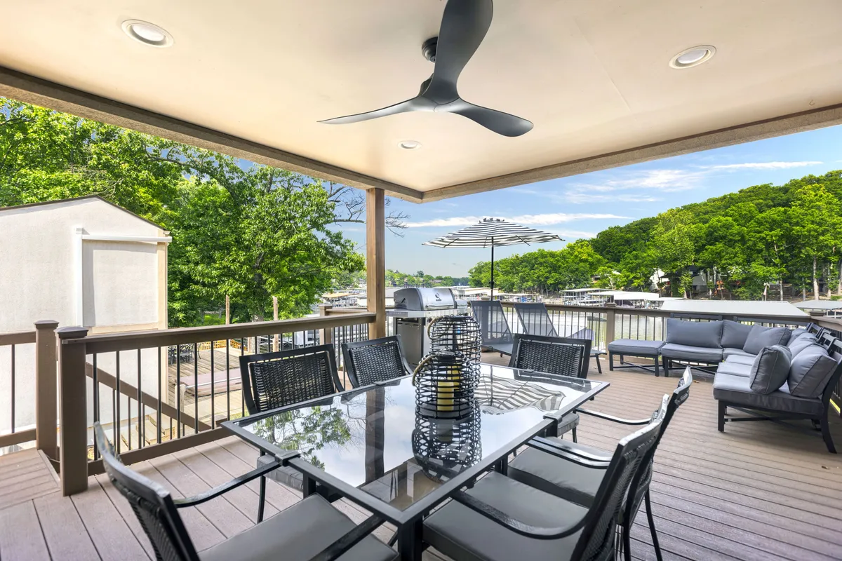 The image showcases a stylish outdoor dining area with a modern table and black chairs, a decorative lantern centerpiece, comfortable seating, a grill for barbecues, and overhead ceiling fan. The area overlooks a beautiful water view, ideal for sunset dining or relaxing outdoors with family and friends.